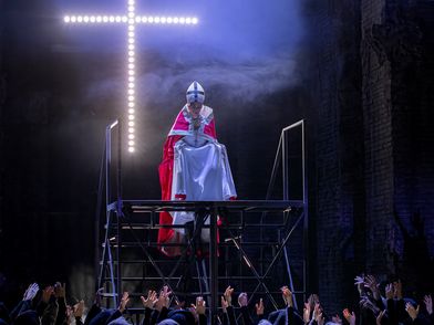 Die Päpstin steht auf einem Gestell, im Hintergrund ein beleuchtetes Kreuz. Am unteren Bildrand recken sich Hände nach oben in Richtung der Päpstin.