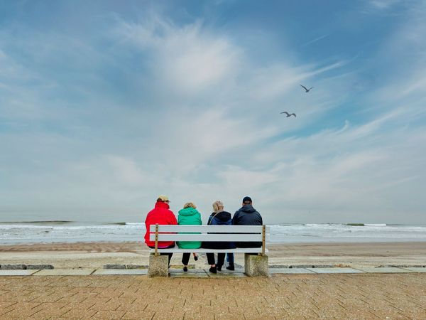 Vier Personen von hinten. Sie sitzen mit bunten Winterjacken auf einer Parkbank an der Standpromenade Norderney. 