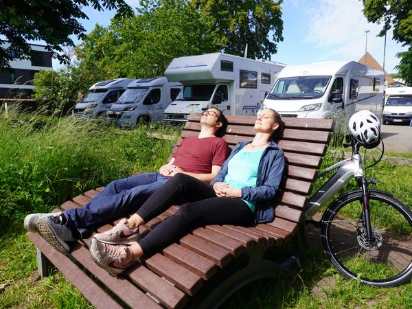 Im Vordergrund liegen ein Mann und eine Frau auf einer Bank in der Sonne, hinter ihnen stehen Wohnmobile auf einem Stellplatz.