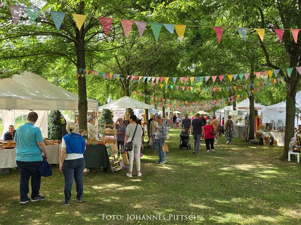 Stände und bunte Girlanden zwischen Bäumen auf einem Sommerfest, mit Besuchern.