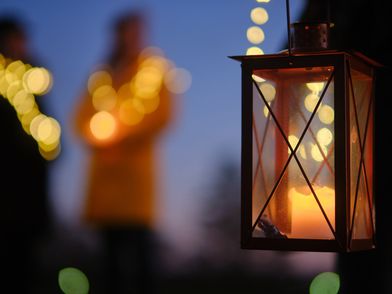 Im Vordergrund eine Laterne mit einer brennenden Kerze, im Hintergrund verschwommene Lichter in der Dunkelheit.