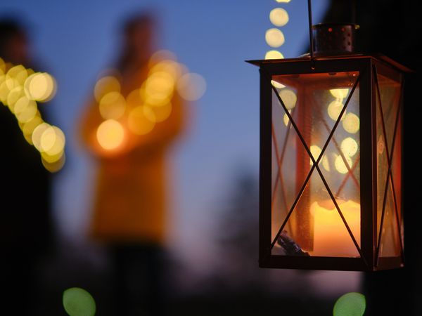 Im Vordergrund eine Laterne mit einer brennenden Kerze, im Hintergrund verschwommene Lichter in der Dunkelheit.